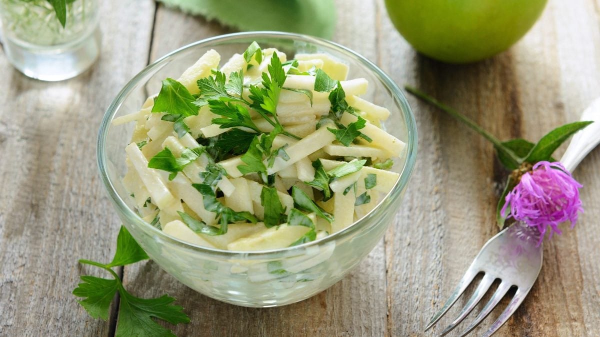 Eine Glasschale mit dem Apfel-Kohlrabisalat auf einem Holztisch mit Petersilie, lila Blüte und Granny Smith Apfel.