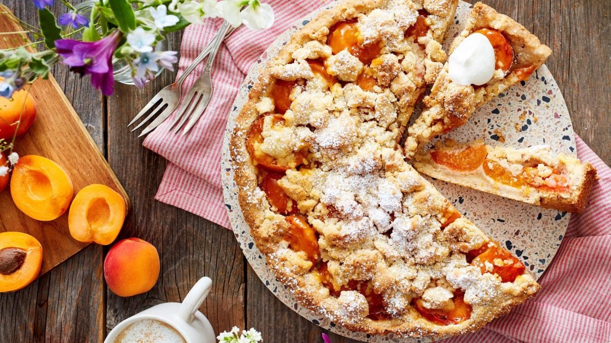 Draufsicht: Angeschnittene Aprikosen-Streusel-Tarte, daneben Aprikosen und Deko auf Holzuntergrund.