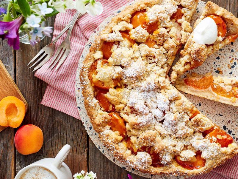Draufsicht: Angeschnittene Aprikosen-Streusel-Tarte, daneben Aprikosen und Deko auf Holzuntergrund.