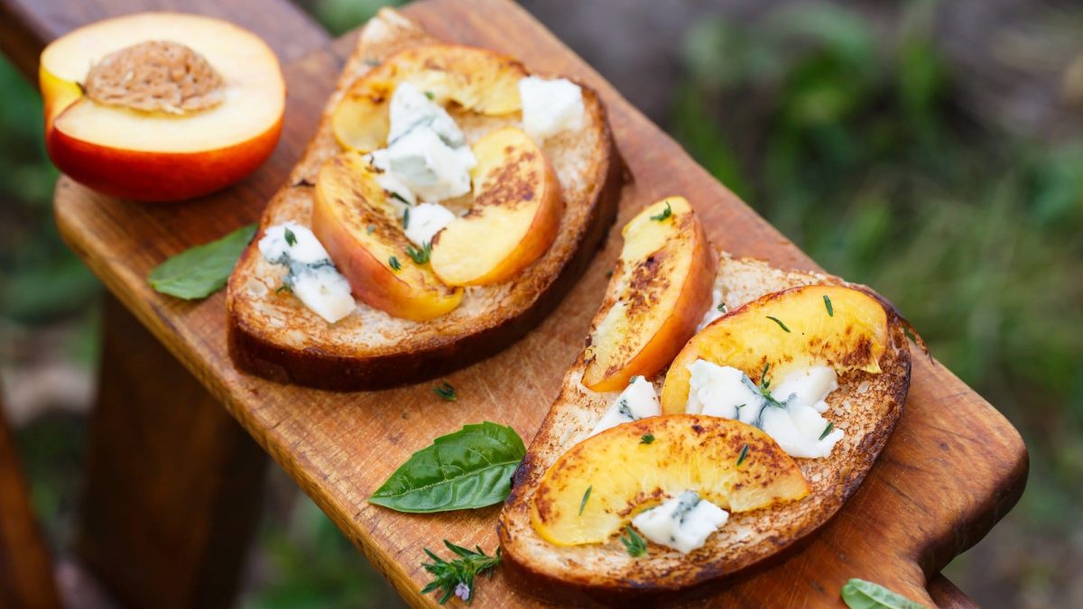 Draufsicht: Zwei Pfirsich-Bruschetta mit Gorgonzola auf einem Holzbrett. Daneben liebe Basilikumblätter und ein halber Pfirsich.