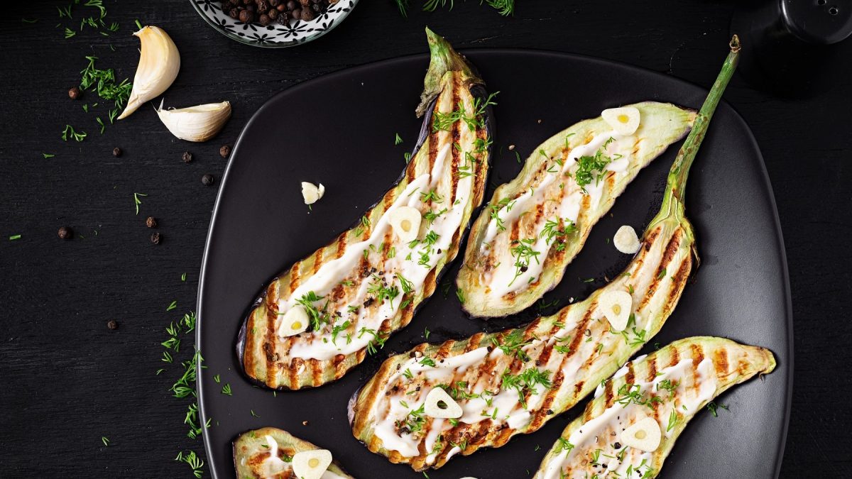 Einige Auberginenschieben mit Grillmuster auf einem schwarzen Teller mit Sesam-Joghurt und frischer Petersilie.