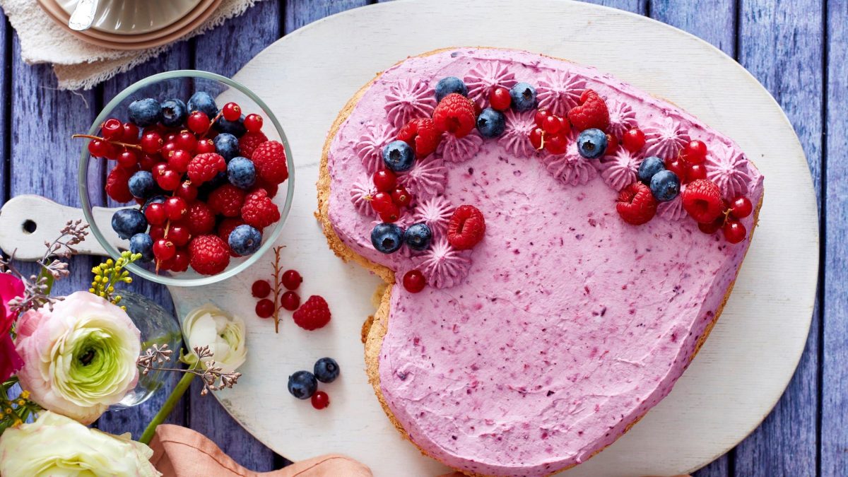 Auf einer Kuchenplatte ist ein Beeren-Quarkherz angerichtet. Es ist mit verschiedenen Beeren dekoriert.