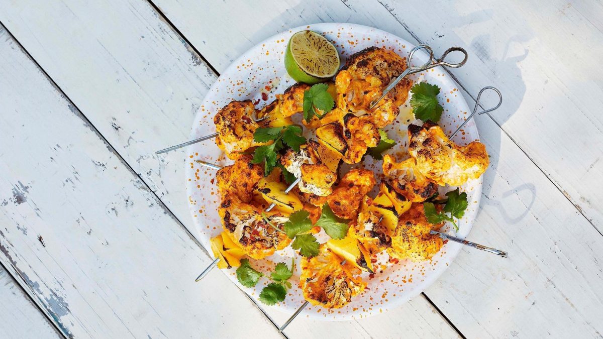 Blumenkohl-Mango-Spieße auf einem Teller mit Limetten und Koriander in der Draufsicht.