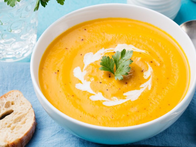 Eine weiße Schüssel mit der Karottensuppe auf einem blauen Untergrund mit weißem Geschirr und dunkelblauer Tischdecke.