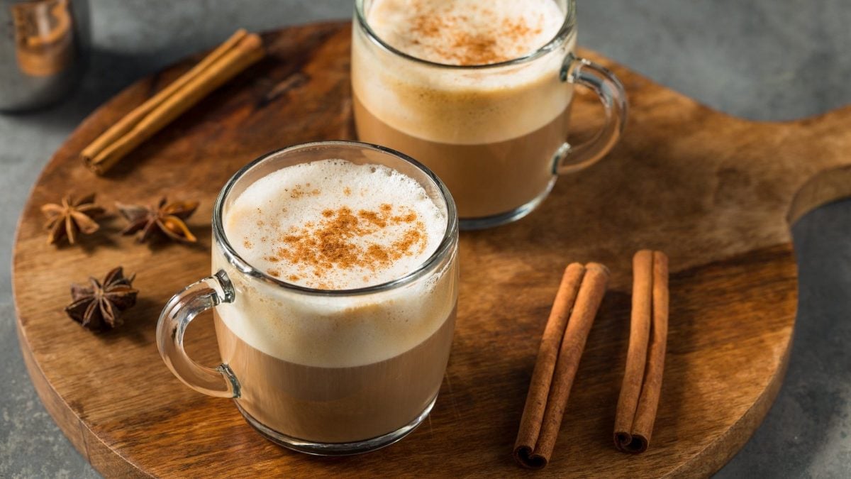 Zwei Gläser Dirty Chai Latte auf einem runden Holzbrett, daneben Zimtstangen und Anissterne.