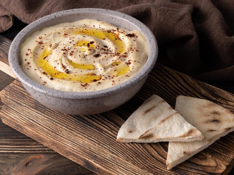 Leicht schräge Draufsicht: Eine runde Schale Hummus und eine ecke abgebrochenes Brot auf Holz.