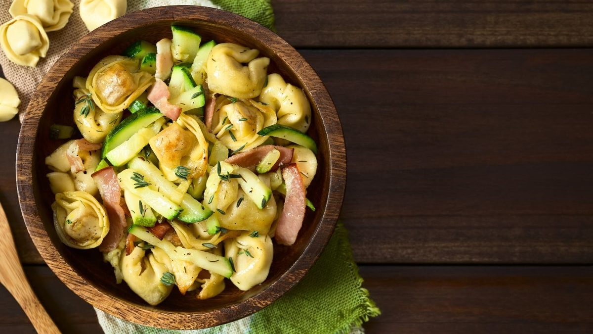 Eine Holzschüssel mit Tortellinisalat mit Pancetta und Zucchini auf einem Holztisch.