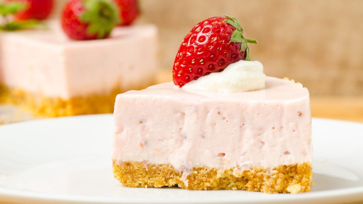 Ein Stück Erdbeer-Cheesecake dekoriert mit einer Erdbeere und einem Klecks Sahne, dahinter sieht man unscharf den angeschnittenen Kuchen.