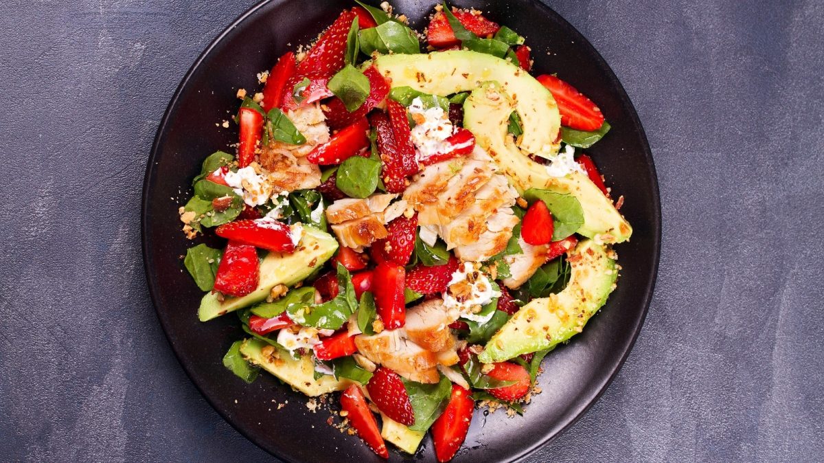 Ein schwarzer Teller mit Erdbeersalat mit Hähnchen und Feta auf einem grauen Tisch.