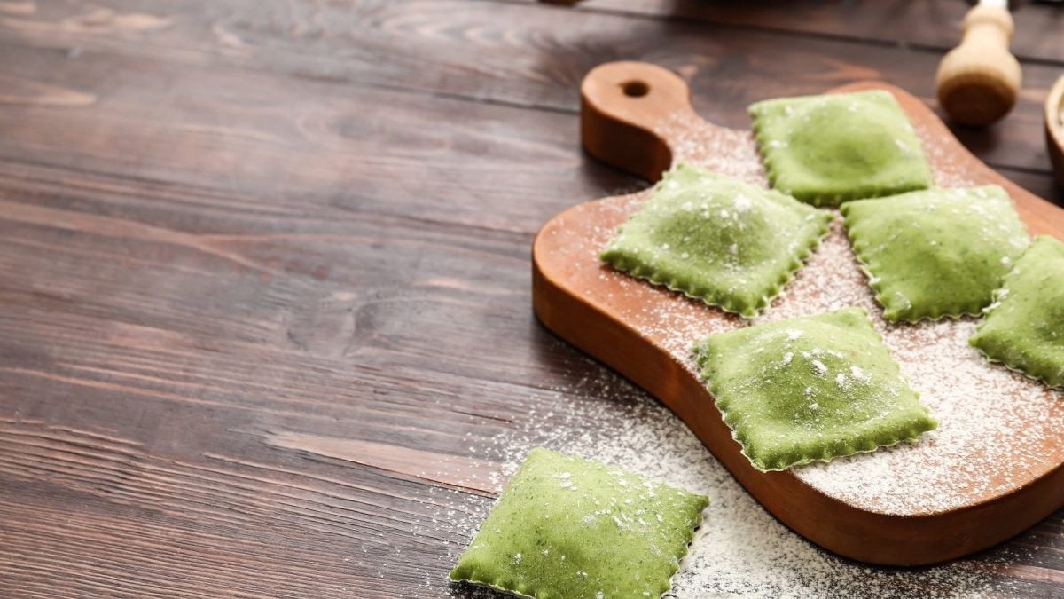 Draufsicht: UNtergrund aus Holz, darauf eine Holzbrettchen mit selbstgemachten Spinat-Ravioli, die noch mit Mehl bestäubt sind.
