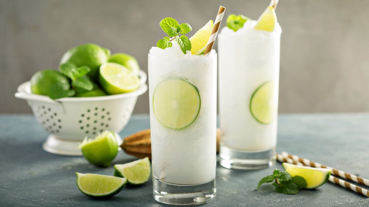 Zwei Gläser Frozen Gin Tonic mit Minze und Limettenspalten garniert, dahinter ein Sieb mit Limetten.