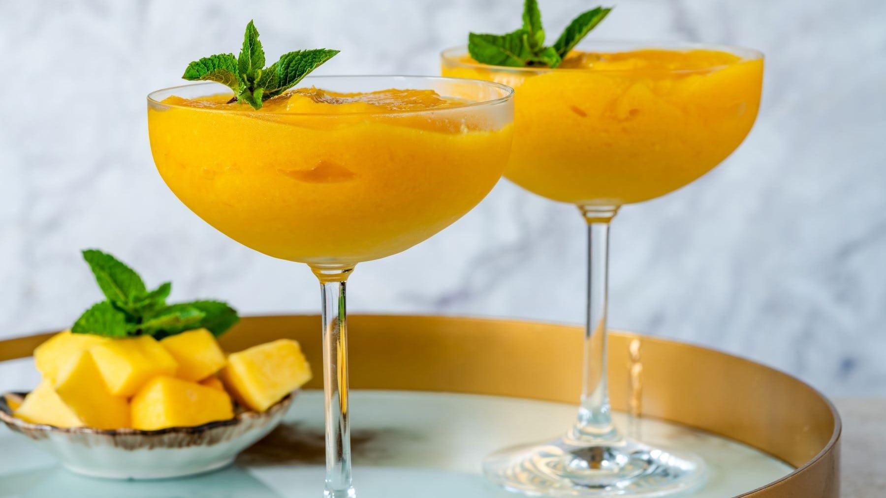 Zwei Frozen Mango Daiquiri mit Minzen auf Tablett. Im Hintergrund eine Schale mit Mangostücken und Minze. Frontalansicht.