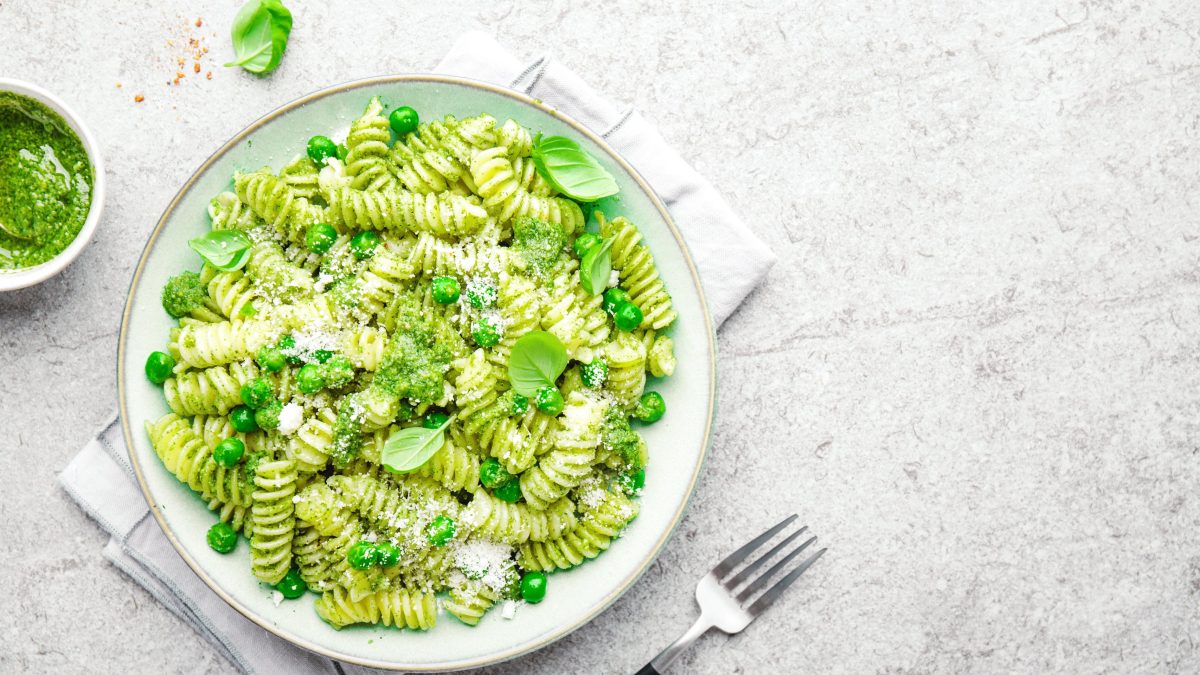 Draufsicht: ein weißer Teller Nudeln mit Basilikum und Erbsen auf einem hellen Untergund. Daneben liege eine Schale mit einem grünen Pesto und eine Gabel.