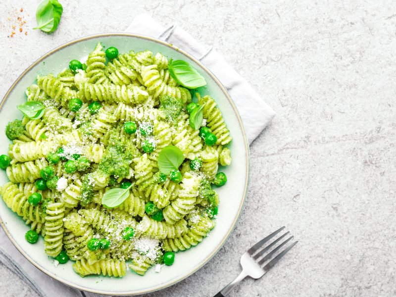 Draufsicht: ein weißer Teller Nudeln mit Basilikum und Erbsen auf einem hellen Untergund. Daneben liege eine Schale mit einem grünen Pesto und eine Gabel.