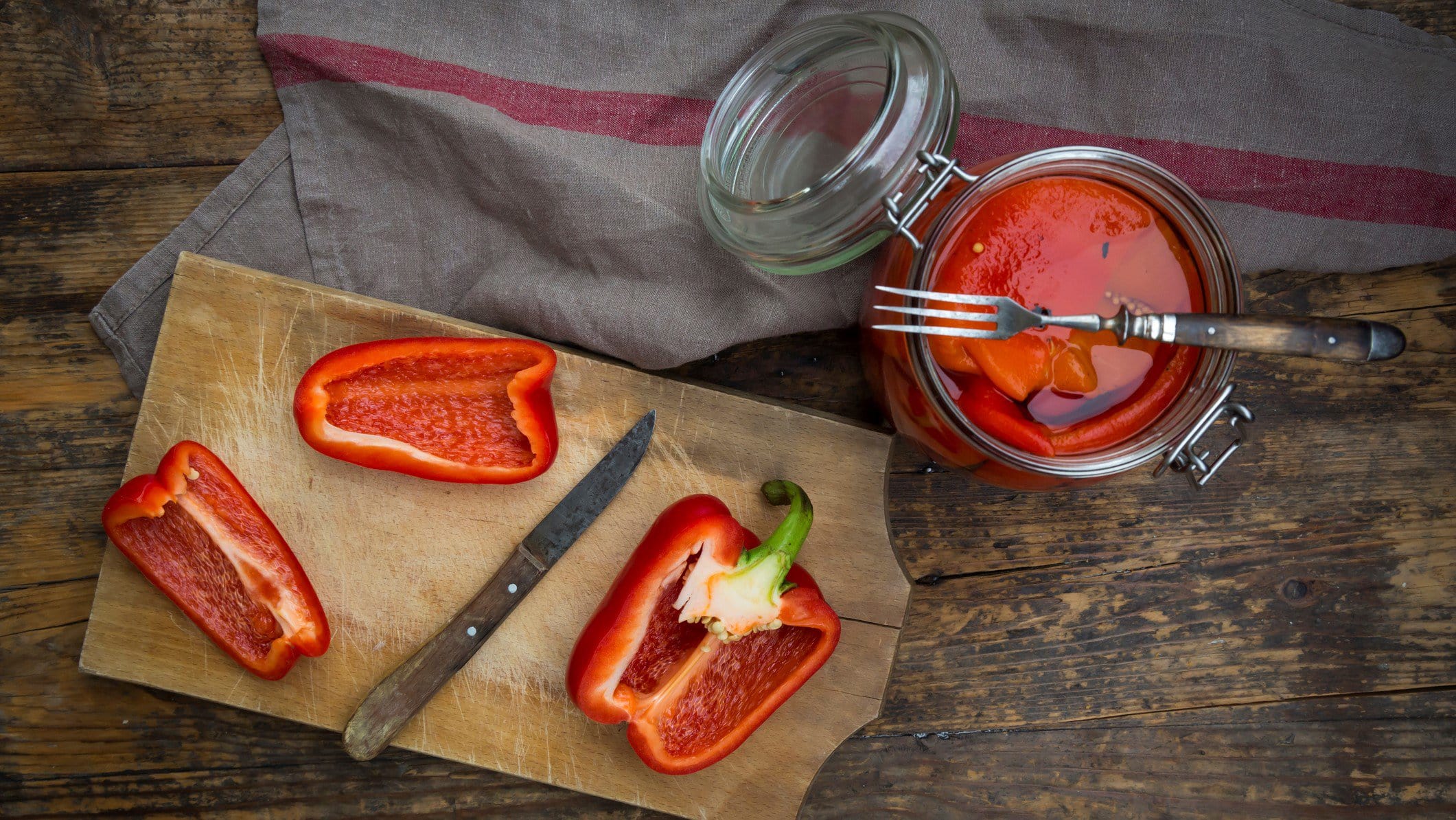 Antipasti geröstete Paprika in Olivenöl in einem Glas mit Gabel, geschnittener Paprika mit Messer auf Holzbrett und grauem Tuch auf Holz. Draufsicht.