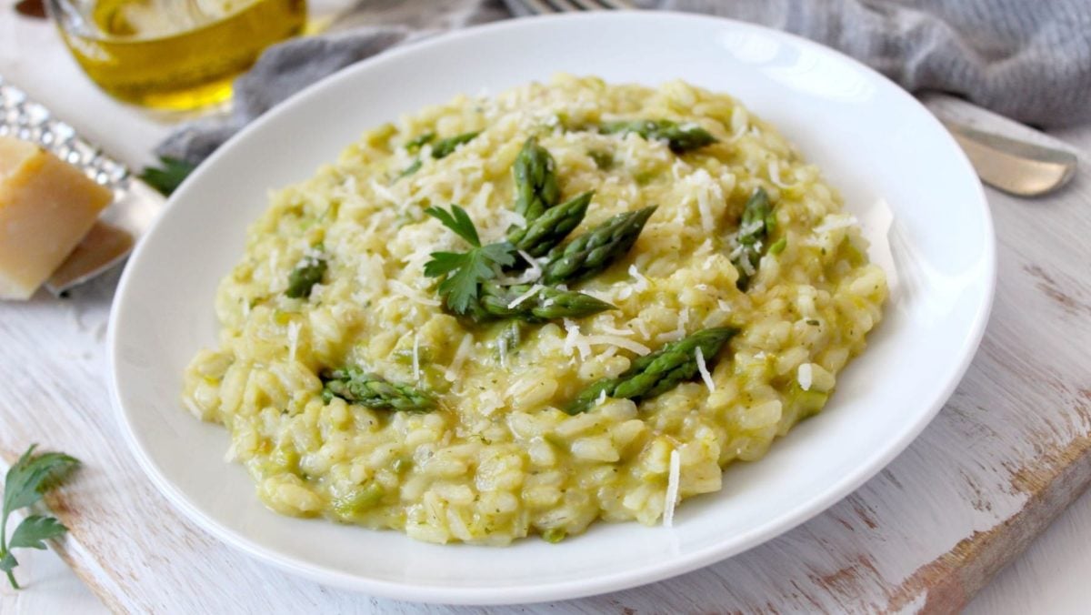 Gorgonzola-Spargel-Risotto auf weißem Teller und hellem Untergrund. Im Bild eine Schale mit Olivenöl und Parmesan auf Käsereibe. Draufsicht.