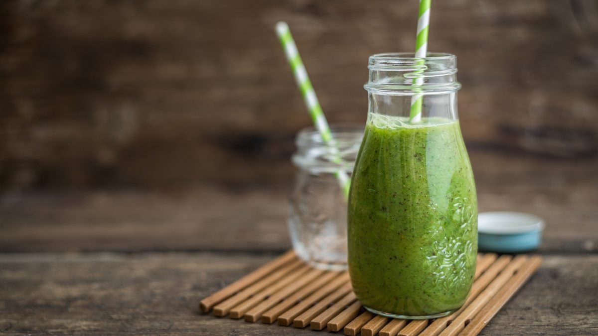 Green Goddess Smoothie im Glas mit grünem Strohhalm auf Bastmatte und Holz. Daneben leeres Glas und Schraubdeckel. Frontalansicht.