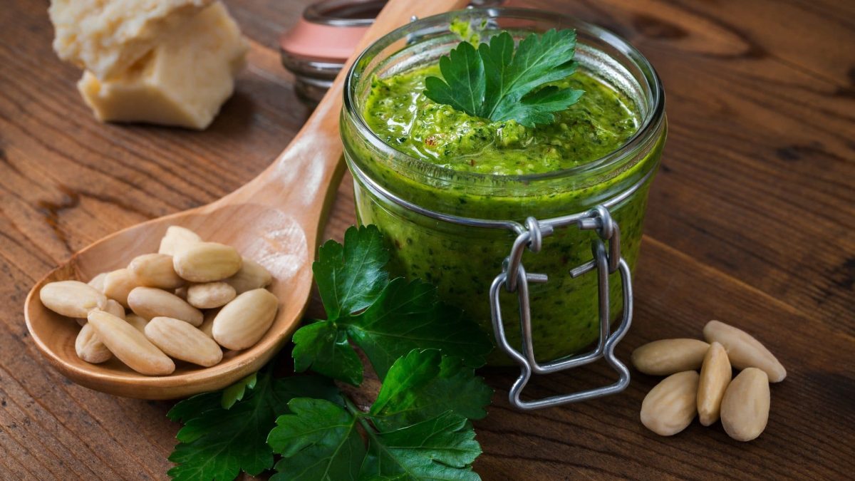 Draufsicht schräg: Auf Holz steht ein Glas Mandel-Petersilien-Pesto. Daneben liegen Mandeln ohne Haut, sowie ein Holzlöffel.