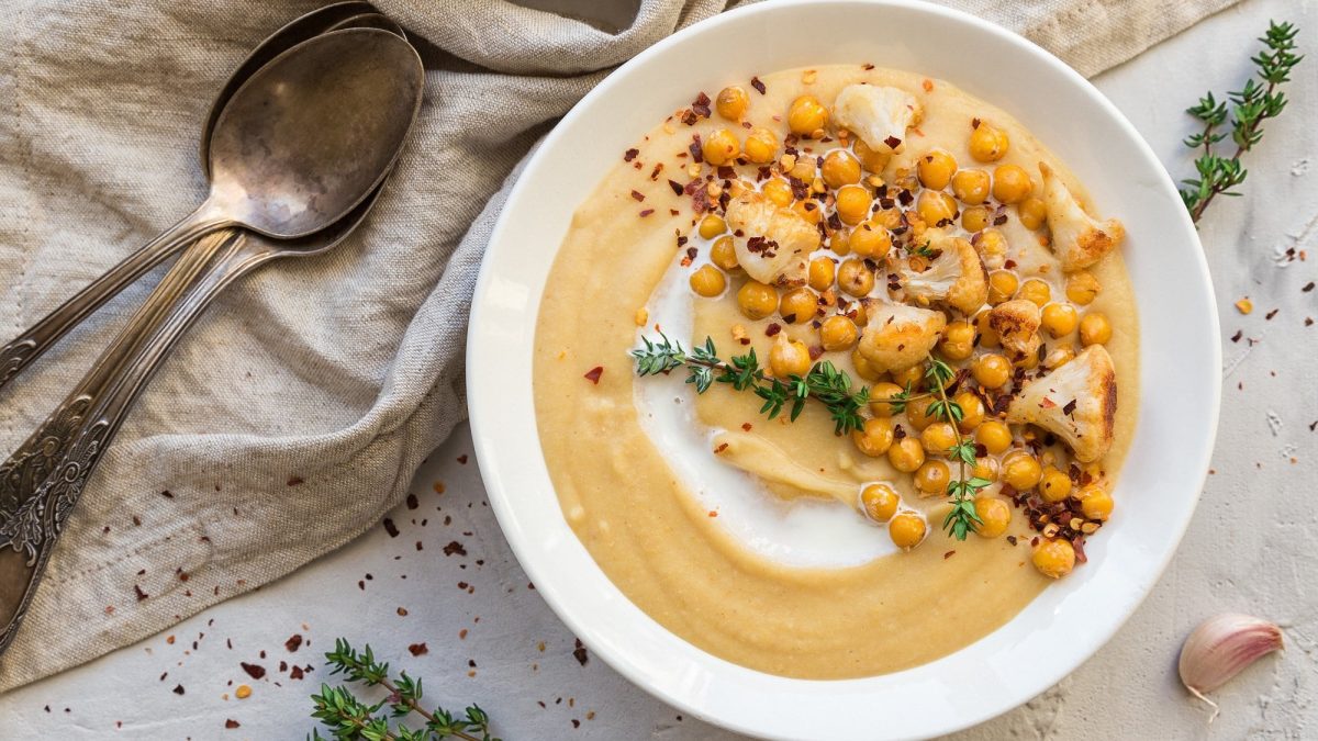 Ein Teller Hummus-Suppe mit Kichererbsen, Pfeffer und Kräutern garniert, Draufsicht, daneben zwei Löffel und Knoblauchzehen.
