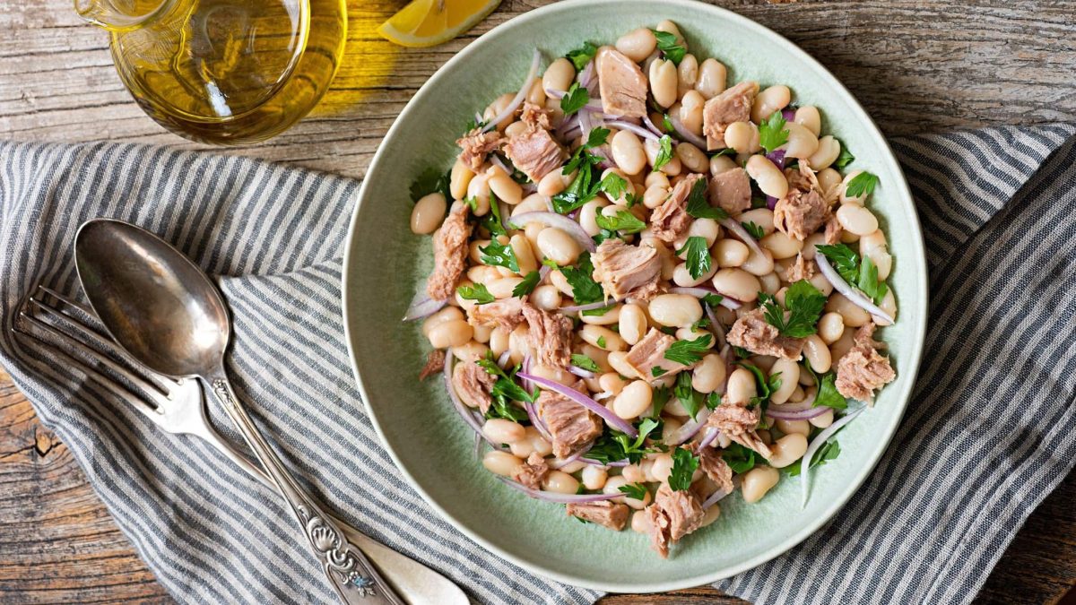 Draufsicht: Auf einem Holztisch steht ein weißer Teller Insalata di fagioli e tonno. Daneben sind ein Besteck, ein Glas mit einer ölig-gelben Flüssigkeit sowie ein Tuch.