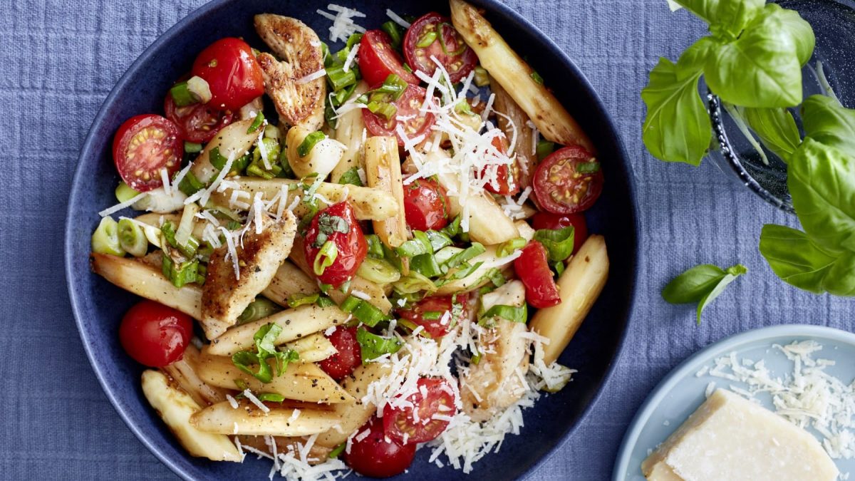 Eine dunkelblaue Bowl mit Spargel, Hähnchenfilet, Tomaten, Parmesan und Frühlingszwiebeln auf einem blauen Hintergrund. Rechts oben ein Basilikum, rechts unten Parmesan.