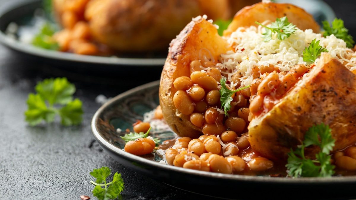 Frontal: Zwei Ofenkartoffeln mit gebackenen Bohnen und Käse liegen auf dunklen tellern auf einem dunklen Untergrund.