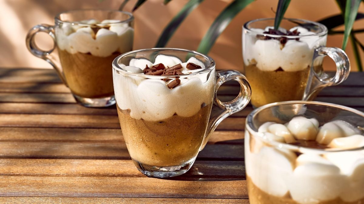 Vier Espressogläser gefüllt mit Kaffeecreme und Sahnehäubchen on top auf einem Holztisch im Halbschatten. Im Hintergrund ein paar Palmenblätter.
