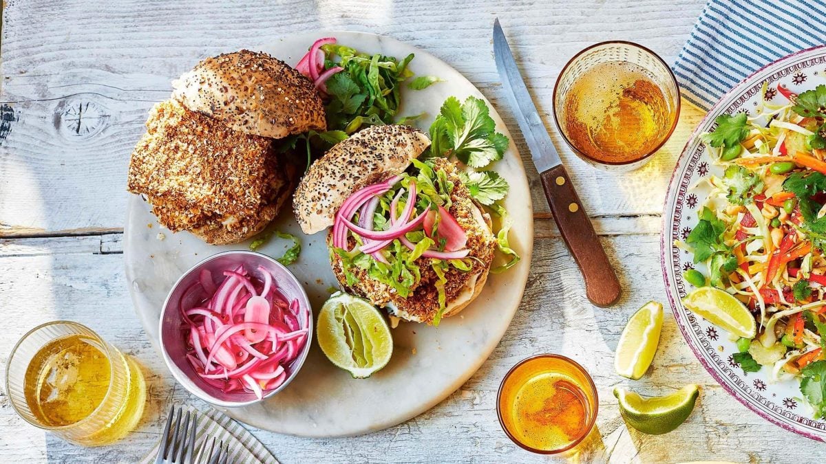 Ein gegrillter Katsu-Burger auf einem Teller mit pinken eingelegten Zwiebeln in der Draufsicht, daneben gefüllte Gläser, ein Messer und eine Schale Salat.