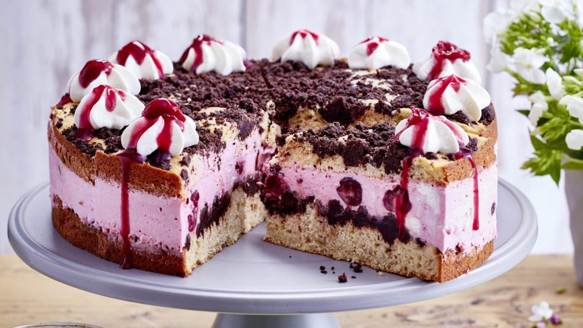 Auf einem Tortenteller steht eine angeschnittene Kiba-Streuseltorte. Sie ist mit Sahnetuffs verziert.