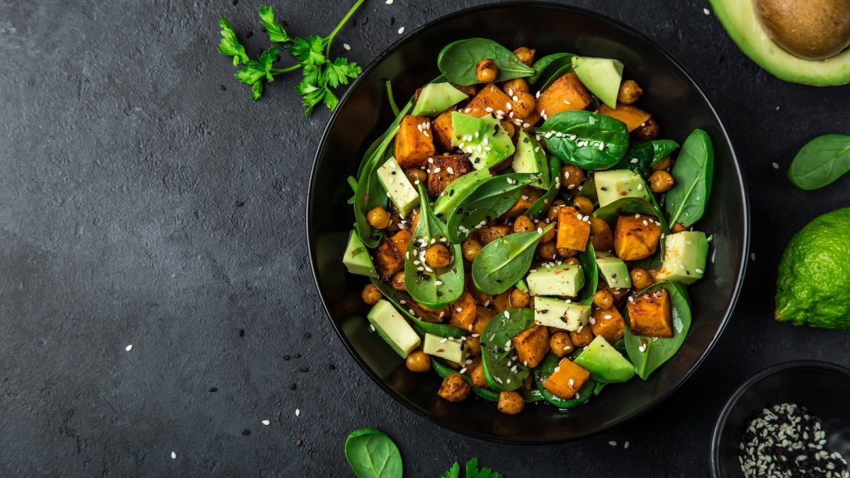 Draufsicht: Auf dunklem Untergrund steht eine schwarze Schüssel Kichererbsen-Spinat-Salat mit Feta.