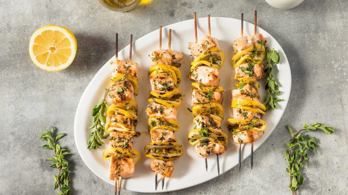 Mehrere Lachs-Kebap mit gelber Zucchini und Zitronenmarinade und Kräutern auf weißem Teller und hellem Untergrund. Daneben Kräuter und Zitrone. Draufsicht.