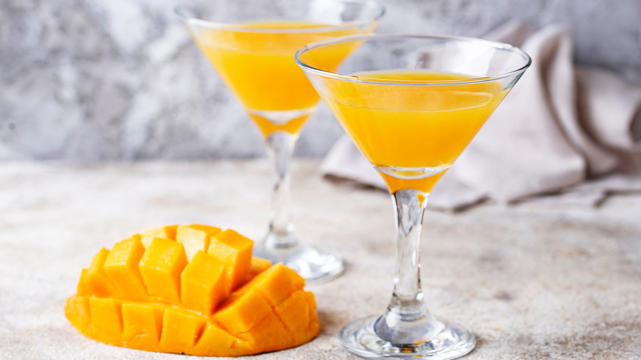 Selbst gemachter Mangolikör in zwei Gläsern mit einer aufgefächert geschnittene Mango auf grauem Untergrund. Frontalansicht.