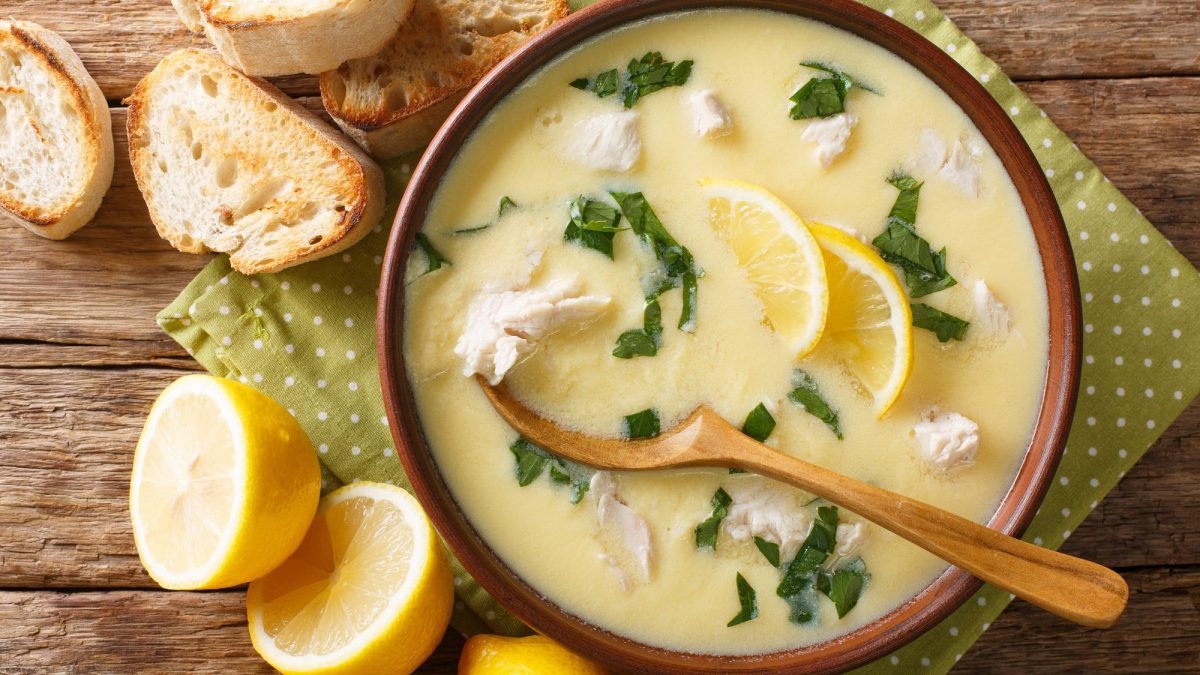 Ein Teller mediterrane Hühnersuppe mit Zitrone und Petersilie auf einem Holztisch. Daneben liegen Zitronenhälften.