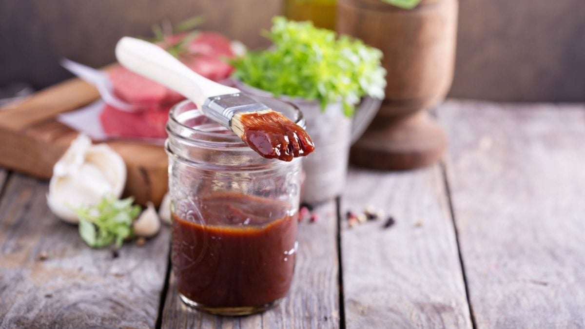 Original Mississippi Barbecue Sauce mit Marinadepinsel, Kräutern, Knoblauch und einem Holzbrett mit Fleisch auf Holt. Frontalansicht.