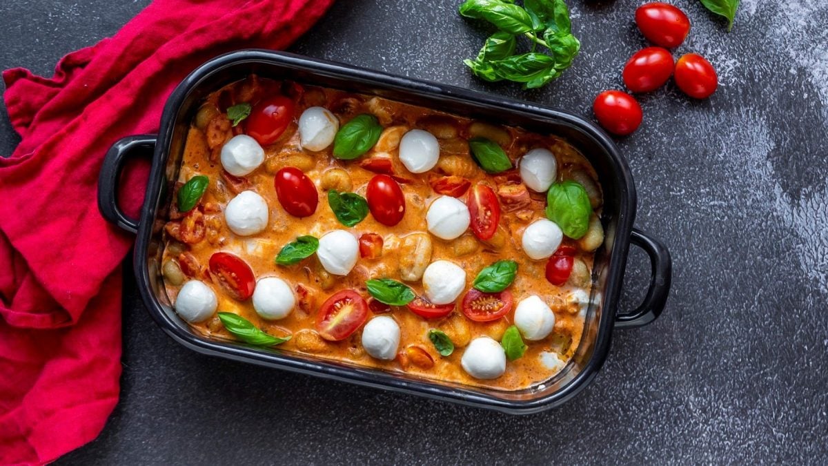 Ofen-Gnocchi mit Tomate & Mozzarella in dunkler Auflaufform. Daneben liegen ein rotes Tuch, Tomaten und einige Blätter Basilikum.