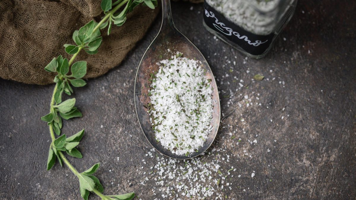 Draufsicht: Ein Löffel selbst gemachtes Oregano-Salz auf einem dunklem Untergrund. Daneben liegt frisches Oregano.