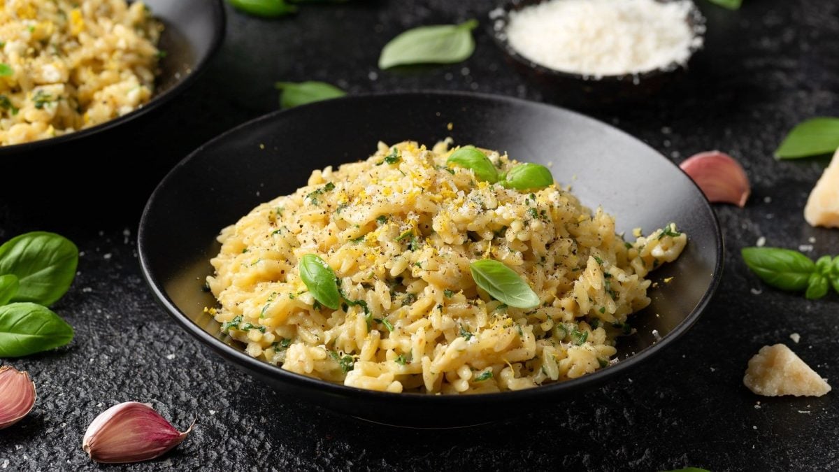Draufsicht: eine Schüssel Orzo al Limone auf einem dunklen Untergrund. Drum herum liegen Basilikumblätter, geriebener Parmesan, und Knoblauchzehen.