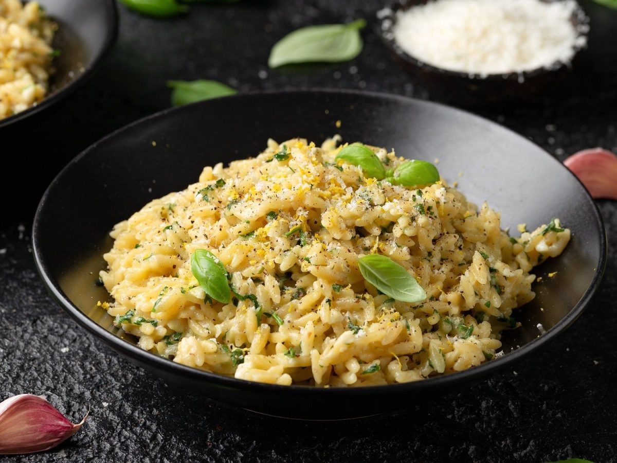 Draufsicht: eine Schüssel Orzo al Limone auf einem dunklen Untergrund. Drum herum liegen Basilikumblätter, geriebener Parmesan, und Knoblauchzehen.