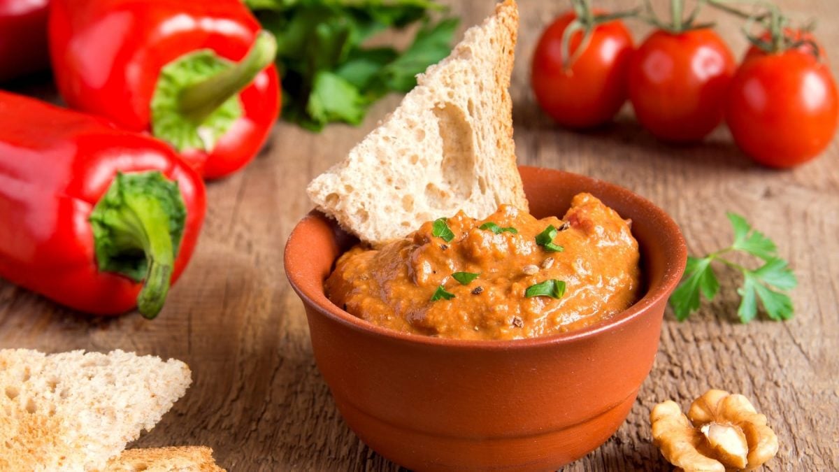 Eine braune Schale mit dem Paprika-Dip auf einem Holztisch. Im Hintergrund Paprika und eine Walnuss.