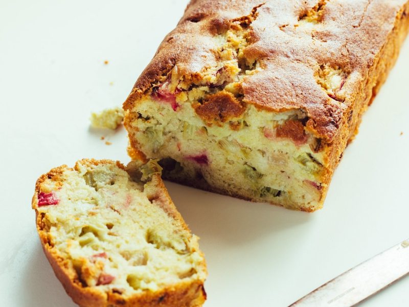 Ein Stück vom Rhabarber-Kastenkuchen udn der Rest davon auf einem hellen Untergrund.