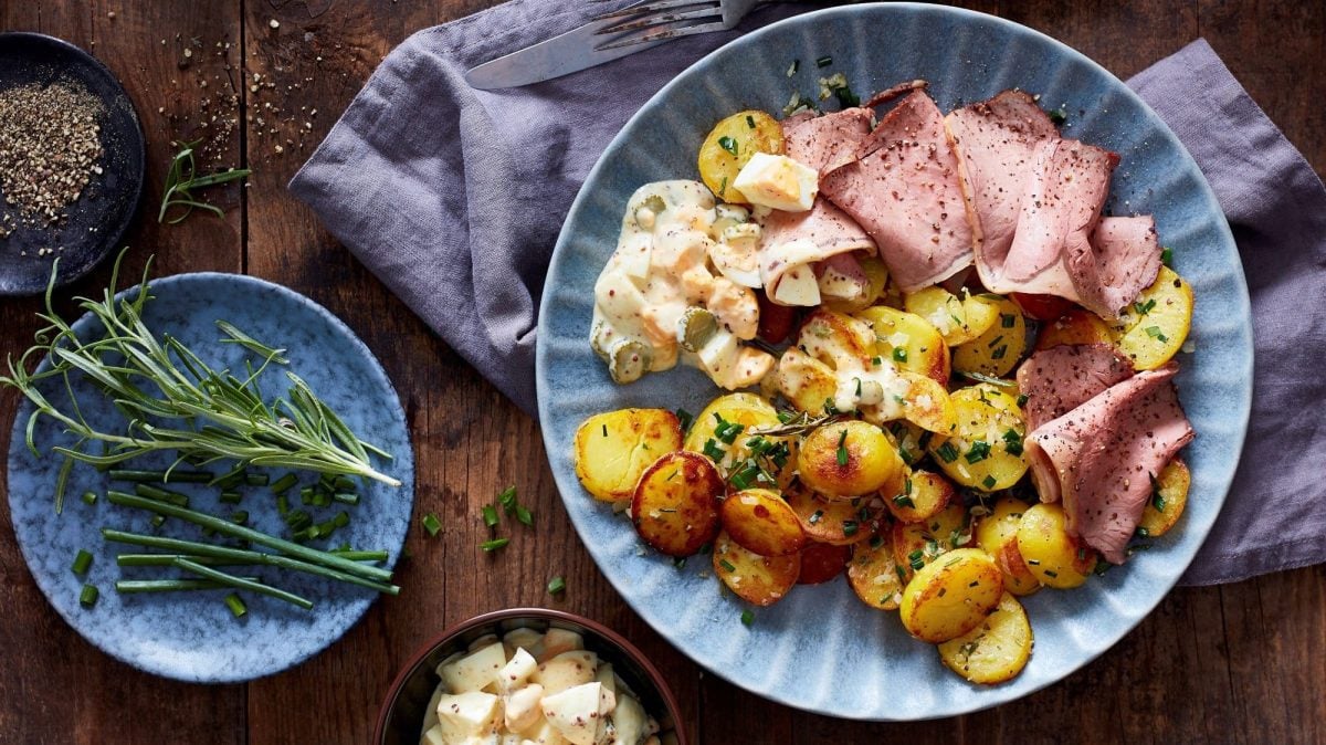 Draufsicht: Ein Teller Kräuter-Kartoffeln mit Roastbeef liegt auf einem dunklen Holztisch. Daneben liegen Rosmarin, kein geschnittenes gekochtes Ei, Pfeffer und ein Tuch.