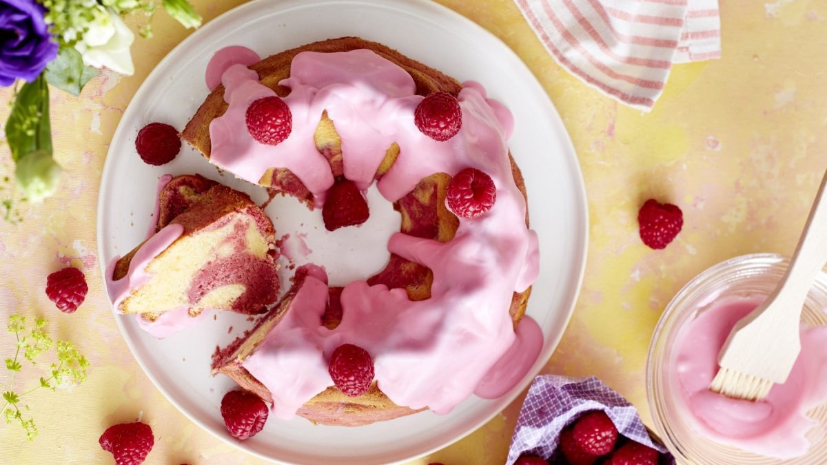 Ein Rosa Marmor-Gugelhupf mit Himbeeren auf einem weißen Teller, auf einem gelben Tischtuch. Rechts unten eine rosa Glasur und vereinzelt ein paar Himbeeren.