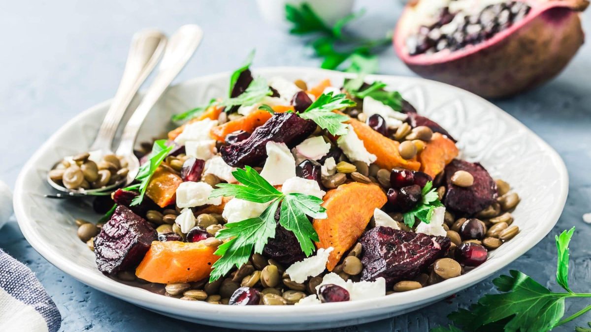 Rote Bete-Linsen-Salat mit Feta, Karotte und Granatapfel mit Besteck auf weißem Teller. Daneben Petersilie und ein halber Granattapfel. Frontalansicht.