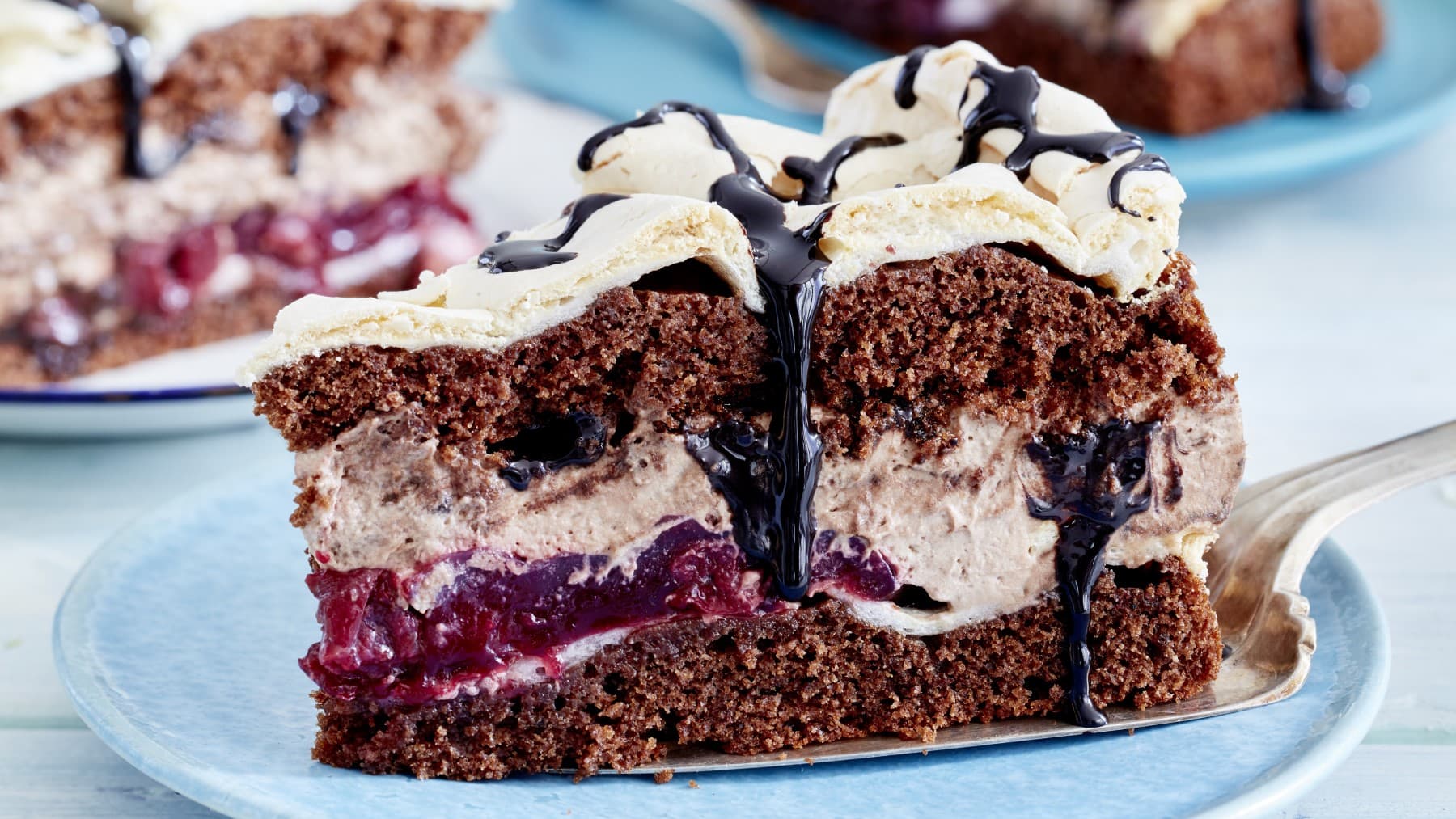 Ein Stück Torte auf einem blauen Teller. Die Torte besteht aus mehreren Schichten. Unten eine Schokoladenkuchenschicht, dann eine Baisermassen, Fruchtschicht, Sahneschicht, Baisermasse, Schokoladenkuchenschicht und noch eine Sahneschicht.