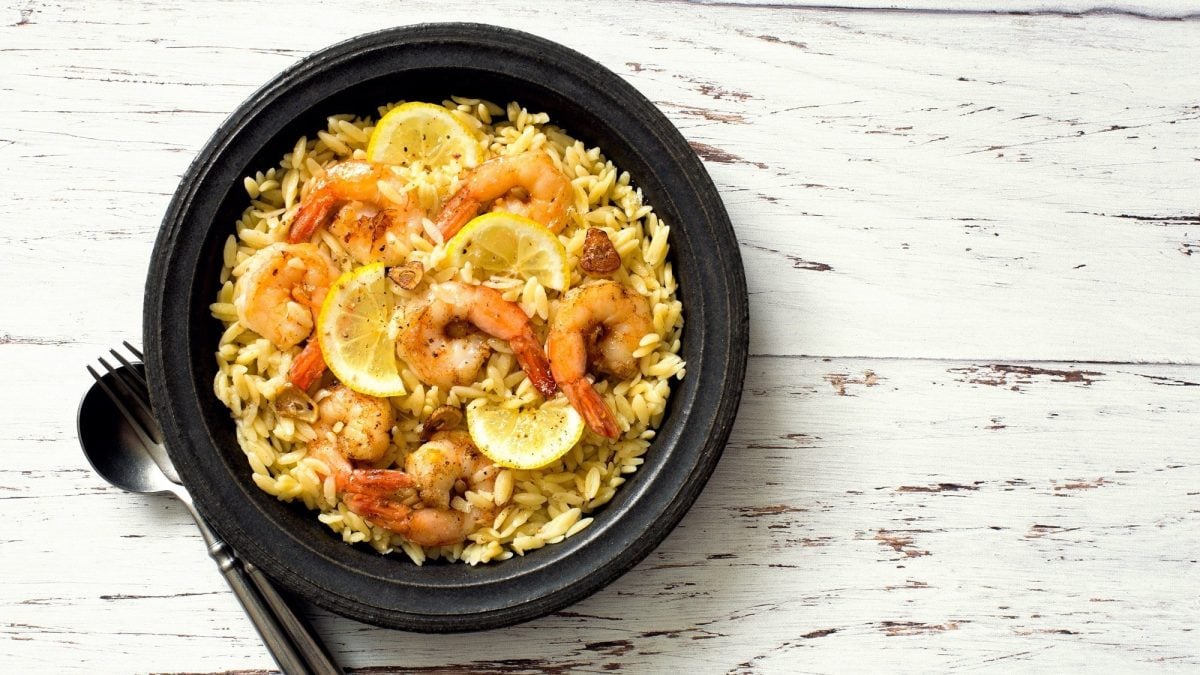 Draufsicht: eine schwarze Schüssel mit Orzo-Garnelen auf einem weißen Untergrund aus Holz: Orzo-Garnelen-Pfanne