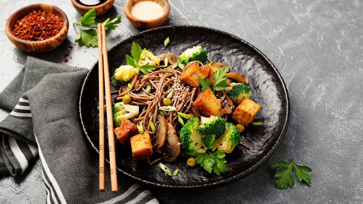 Ein schwarzer Teller mit Soba-Nudelsalat auf einem grauen Steinuntergrund.
