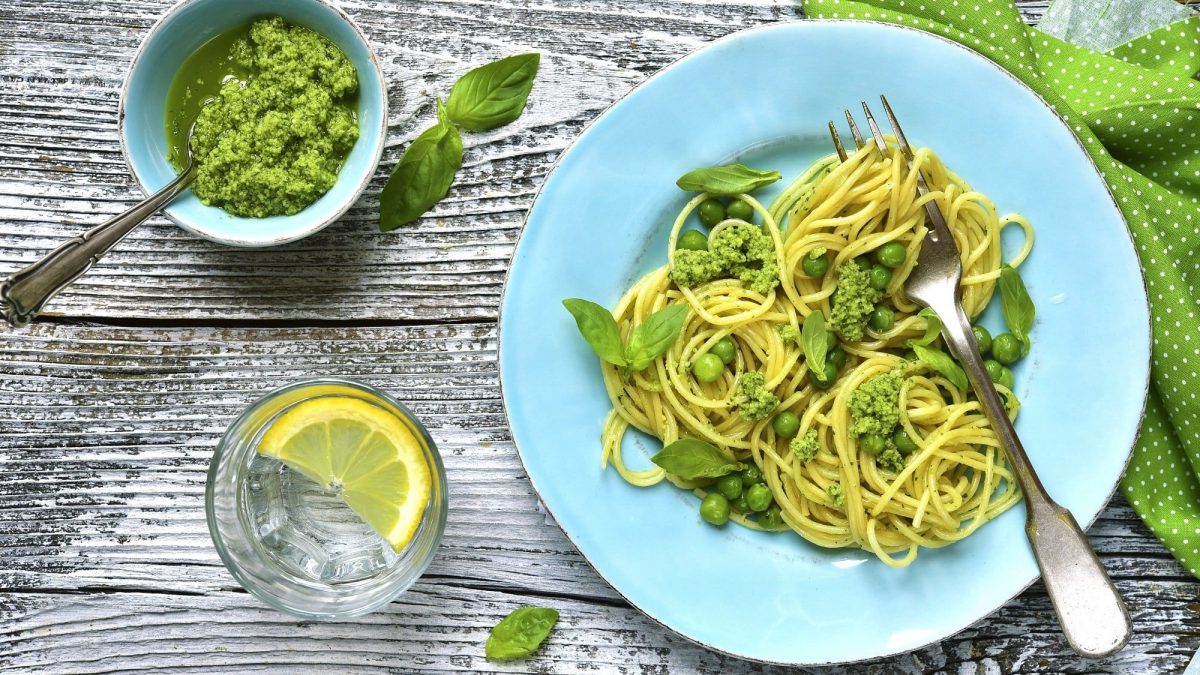 Draufsicht: ein hellblauer Teller mit Spaghettini mit Erbsen und Pesto auf einem Holztisch. Daneben stehen ein Glas Zitronenwasser und eine Schale mit Pesto.