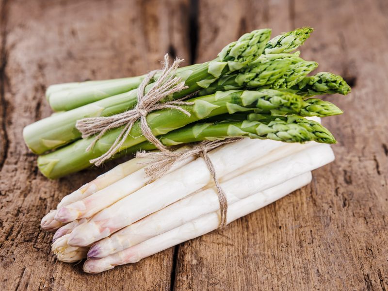 Zwei Bündel grüner und weißer Spargel, mit Bast umwickelt, liegen nebeneinander auf rustikalem Holzuntergrund.