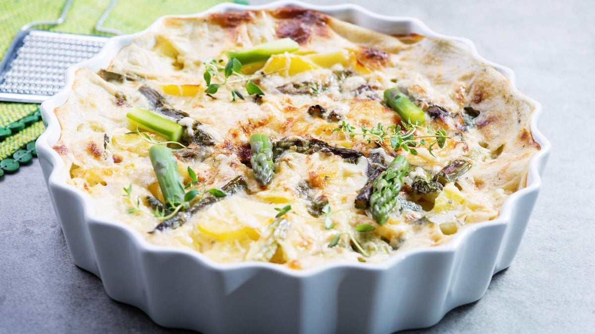 Spargel-Kartoffel-Gratin mit Parmesankruste und mediterranen Kräutern in weißer Kasserole auf grauem Untergrund mit Kräutern. Draufsicht.