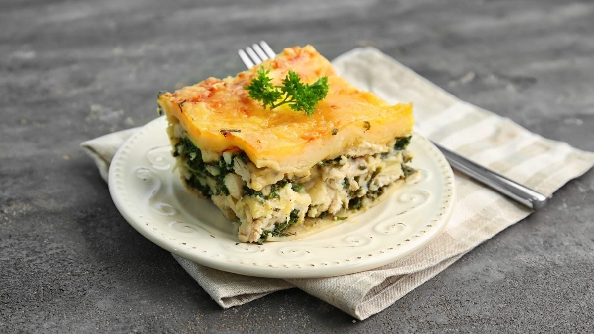 Ein weißer Teller mit einem Stück der Spinat-Lasagne auf grauem Untergrund mit einer hellen Serviette und silbernen Gabel.